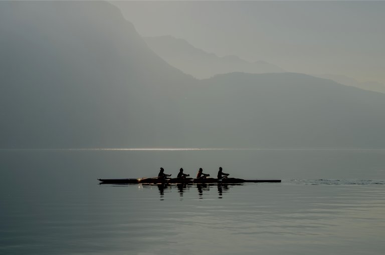 Four people are rowing a boat on a calm body of water with mist-covered mountains in the background. Their synchronized teamwork, combined with the soft lighting and fog, creates a serene and peaceful atmosphere.