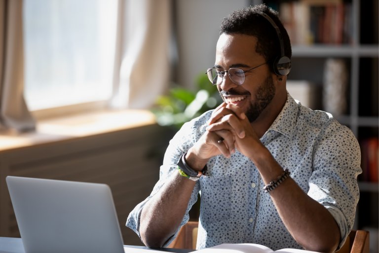 A person wearing glasses and headphones is sitting at a desk, smiling and looking at a laptop screen. They are clasping their hands together in front of them. In the background, there are bookshelves and a window with sunlight streaming in, creating an inviting workspace for remote employees.