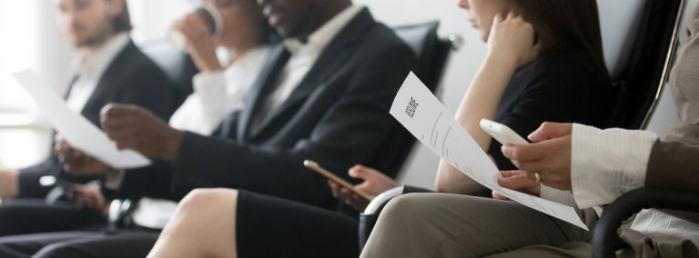 Four people sit in a row in what appears to be a waiting area or interview room. Dressed in business attire and holding resumes, they seem focused and engaged. Two of them are also using smartphones, possibly researching Government Jobs or careers in government.