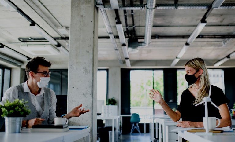 Two people wearing face masks sit at separate desks in a modern office space with exposed pipes and concrete walls. They are engaged in a conversation about careers in government, gesturing with their hands, as natural light streams in through large windows in the background.