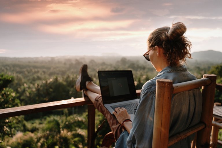 A person with a bun hairstyle is sitting on a wooden chair, working on a laptop. They overlook a lush, green valley as the sun sets, filling the sky with soft colors. The scene exudes tranquility and focus, embodying the essence of remote work in nature's embrace.
