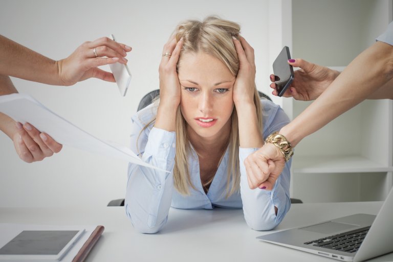 A stressed woman sitting at a desk with her head in her hands is surrounded by multiple hands holding a phone, papers, and a tablet, indicative of the high-pressure demands often associated with medical care.