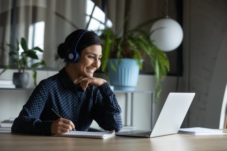 A person wearing headphones is sitting at a table, smiling and taking notes while looking at a laptop. Engaged in remote working, the scene includes a large potted plant and light curtains in the background.