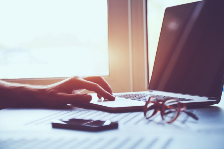 A person's hand is typing on a laptop keyboard near a window with sunlight streaming through, possibly crafting a detailed job description. A pair of eyeglasses and a smartphone are placed on the table next to the laptop, enhancing the warm and cozy ambiance of the scene.