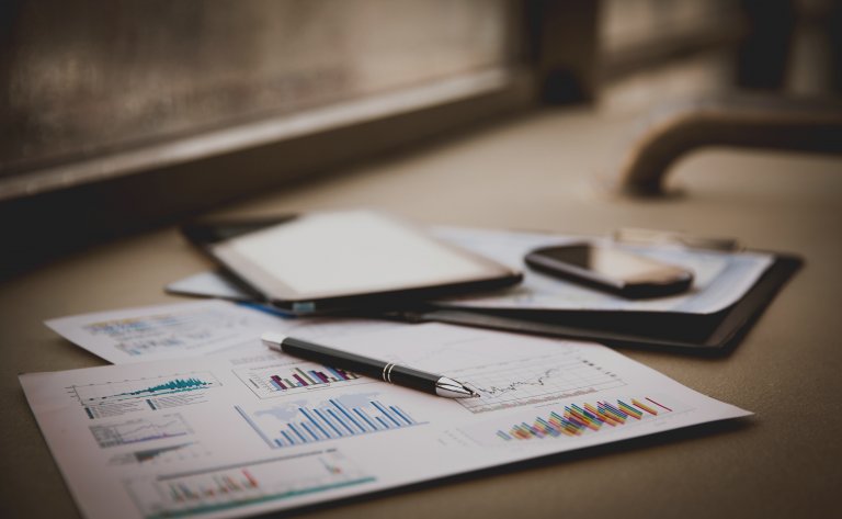 A desk with financial charts and graphs on papers, a pen, a tablet, and a smartphone placed on top, set near a window. The colorful bar and line graphs indicate accounting finance analysis in progress.