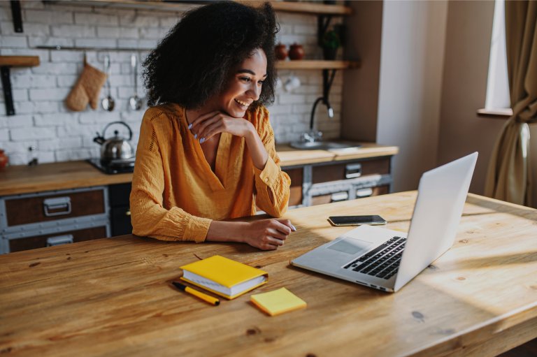 A person with curly hair wearing a yellow blouse is sitting at a wooden table in a kitchen, smiling and looking at a laptop. Yellow notepads, a smartphone, and other essentials for remote work are on the table. Shelves with kitchen items are in the background.
