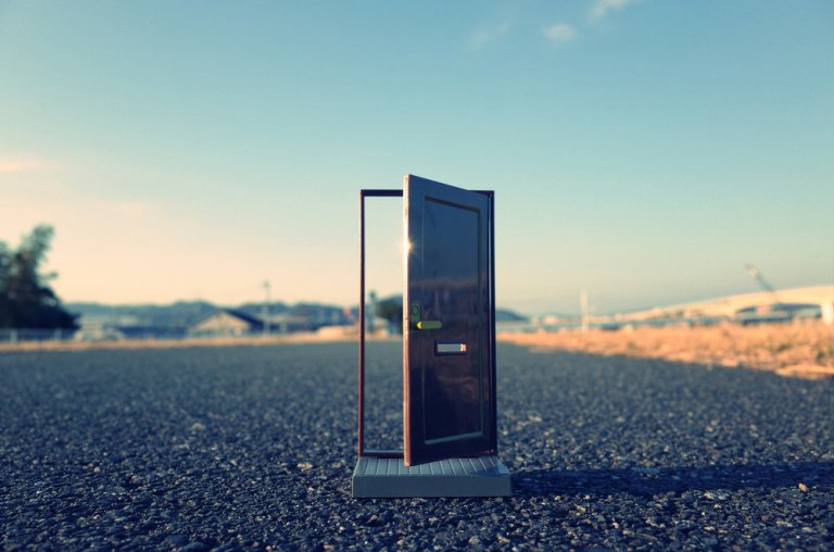A dark brown door stands ajar in the middle of an empty road, leading into an open, expansive landscape under a clear blue sky. The surreal placement of the door contrasts with the natural surroundings.
