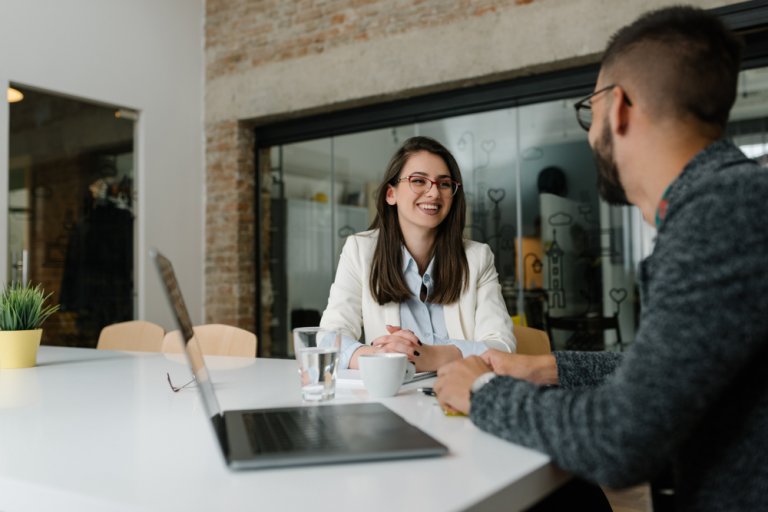 A woman in a white blazer sits at a table, smiling and talking to a man with glasses. They are inside a modern office space with a brick wall and glass partition. A laptop, a cup of coffee, and a glass of water are on the table as they discuss job hunting strategies.
