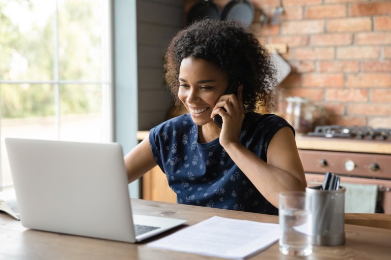 A woman with curly hair smiles while talking on the phone and working on a laptop at a wooden table. She sits in a cozy kitchen with brick walls, a glass of water, and some documents and pens next to her, embracing remote working as natural light filters through a nearby window.