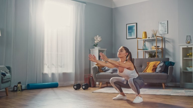 A woman in athletic wear performs a squat exercise in her bright, modern living room, which doubles as a home gym with a sofa, bookshelf, and fitness equipment like a yoga mat and dumbbells. Sunlight streams through the window, creating a warm and energized atmosphere.