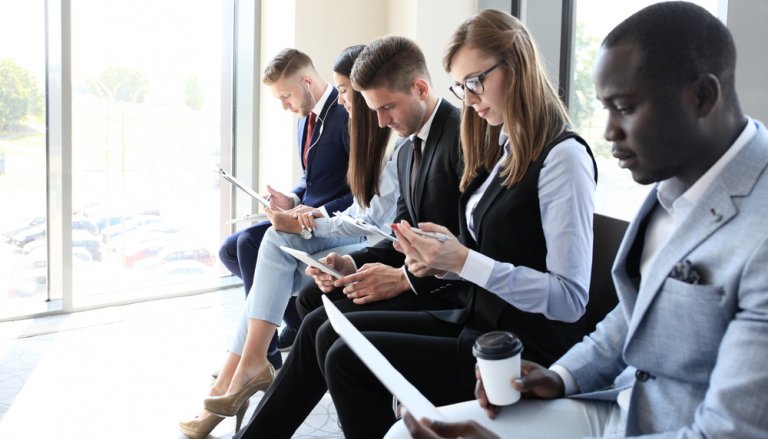 A group of five professionally dressed people sit in a row holding documents and electronic devices, possibly awaiting an interview for a government job in an office with large windows. They appear focused, with one person holding a coffee cup.