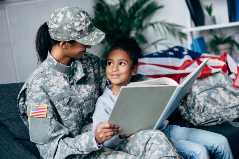 A military personnel in uniform, possibly from ROTC, sits beside a young child on a couch, holding a large book. The child looks up at the military personnel with a smile. An American flag and military gear are visible in the background.