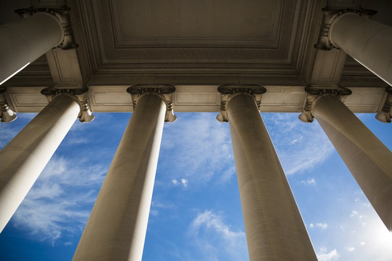 A low-angle view of tall, white columns supporting a classical government building. The sky is bright blue with scattered clouds, providing a striking contrast to the architectural structure. The image captures the grandeur and symmetry of the columns.