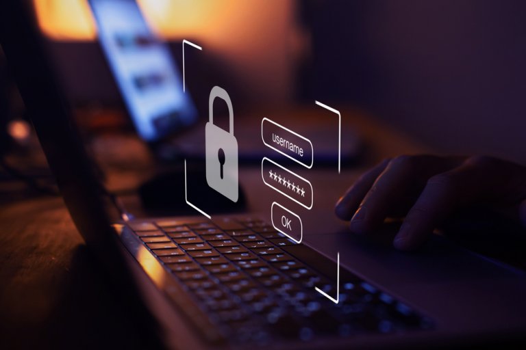 A person uses a laptop in a dimly lit room. A holographic image of a login screen with fields for username and password, a padlock icon, and an "OK" button is superimposed over the keyboard, symbolizing password security and login authentication.
