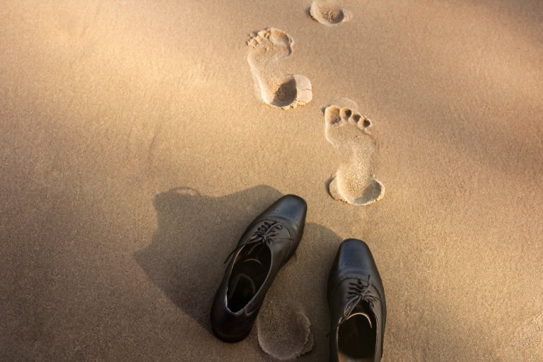 A pair of black dress shoes sit on the sand with barefoot footprints leading away in a quiet, symbolic act of resignation. The sandy beach appears smooth and sunlit, with shadows casting from the shoes.