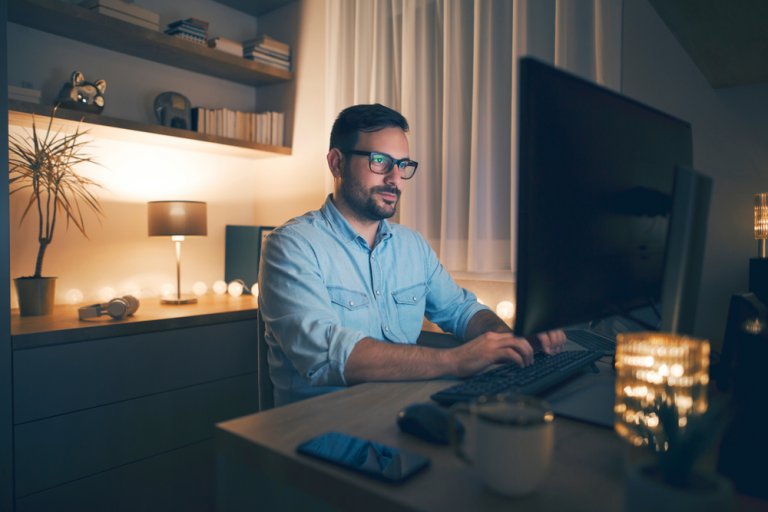 A man wearing glasses is sitting at a desk, typing on a computer keyboard in a dimly lit room, embracing the essence of remote work. The desk is decorated with small lights, a lamp, a potted plant, and a few books. A smartphone and a mug are also placed on the desk.