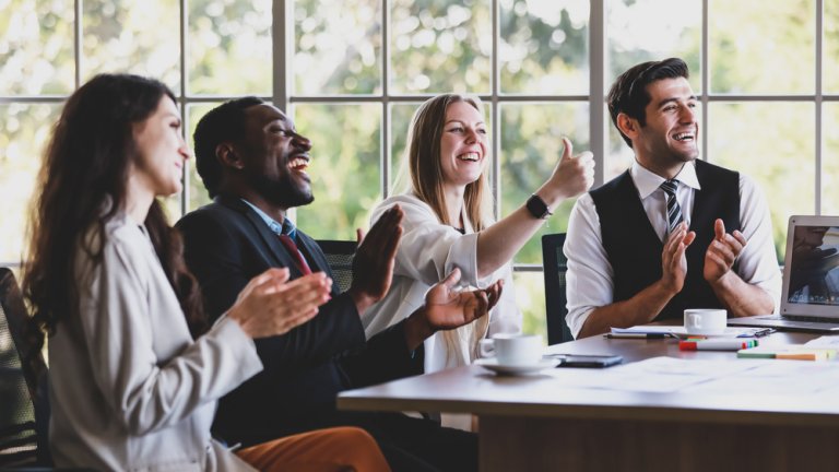 A diverse team of four professionals is sitting at a conference table, smiling and applauding. One woman is giving a thumbs up. The background shows large windows with a view of trees, suggesting a bright and positive atmosphere in a modern office setting.