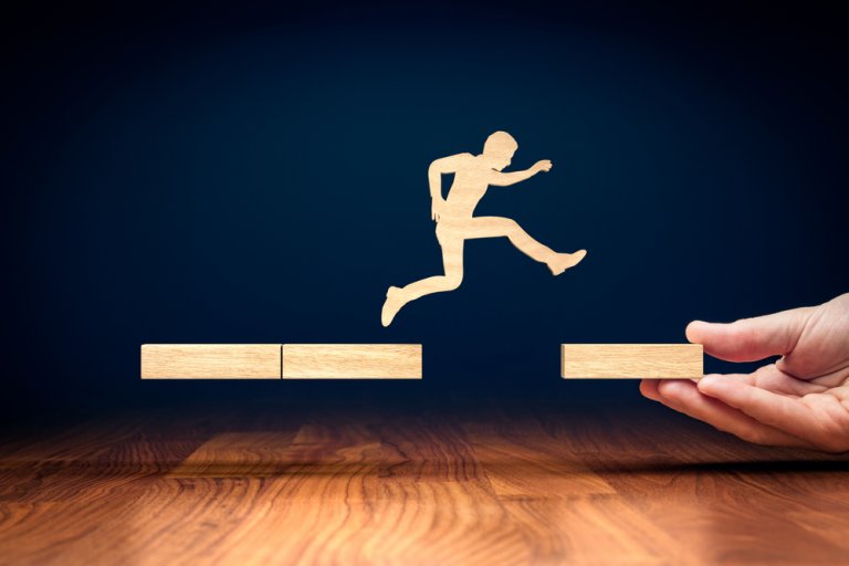 A wooden figure of a person is depicted leaping across a gap between two platforms made of wooden blocks. One platform, symbolizing government support, is being held up by a hand, representing taking a leap or overcoming an obstacle. The dark background creates a stark contrast.
