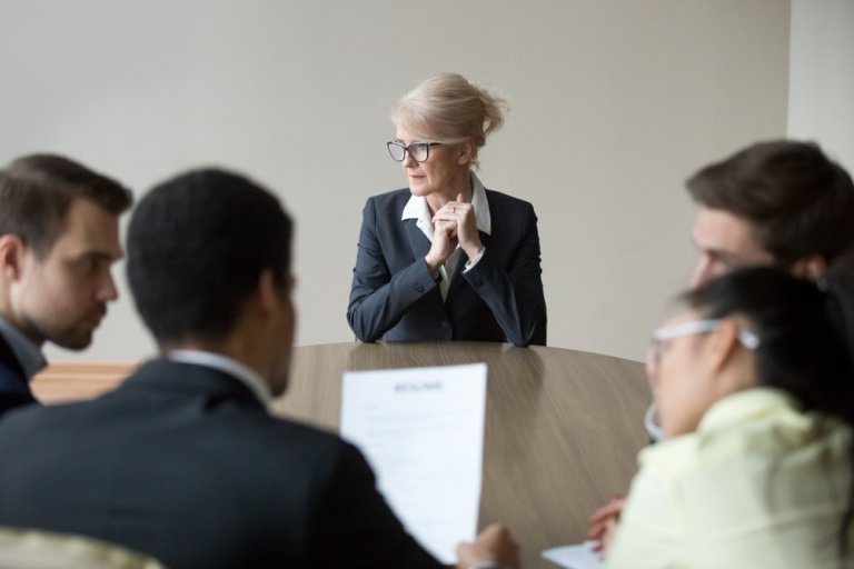 A businesswoman in a black suit and glasses sits at a round table, clasping her hands and looking thoughtful. Surrounding her are four other people, two men and two women, focused on documents in front of them during a meeting within the minimalist room, as if reviewing resumes.