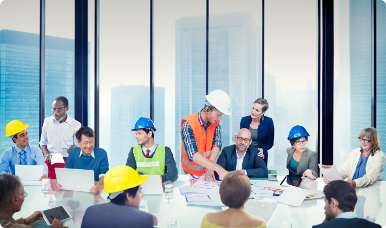A diverse group of professionals, including construction workers wearing hard hats and office staff, gather around a table in a modern conference room with large windows overlooking a cityscape, collaborating on a project with blueprints and laptops.