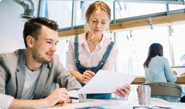 Two people are collaborating in a bright, modern office. A man in a suit jacket sits and looks at a document while a woman in a white shirt and denim overalls stands next to him, holding the paper and pointing at something. Another person works at a desk in the background.