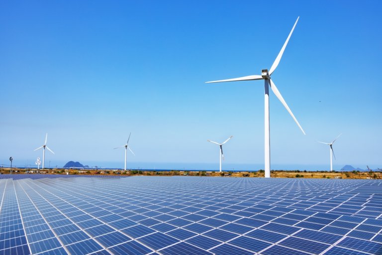 An expansive field of solar panels under a clear blue sky, with several large wind turbines standing tall in the background. The scene is set against distant hills and the ocean, showcasing government investments in renewable energy sources amidst a serene landscape.