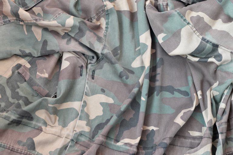 A close-up image of a crumpled, green and brown camouflage-patterned fabric. The fabric appears to be part of a military or outdoor garment, such as a jacket or pants, commonly worn by veterans, featuring various shades of green, brown, and beige in a camouflage design.