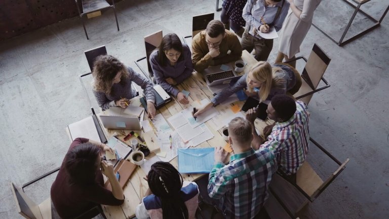 A group of diverse people sit around a large table, embodying DEI principles as they engage in discussion and work. Papers, laptops, and stationery are spread across the table. The setting appears to be a collaborative office or classroom environment.