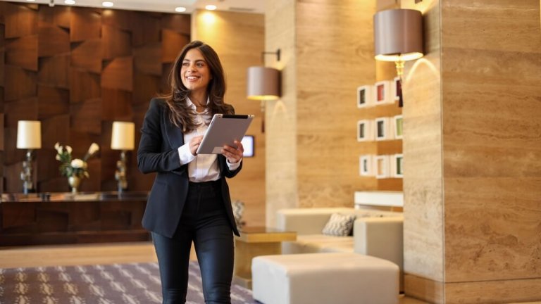 A woman in business attire walks confidently through a modern, stylish lobby, holding a tablet. The lobby features warm lighting, wooden panels, and contemporary decor. She exudes an air of management expertise, appearing focused and professional.