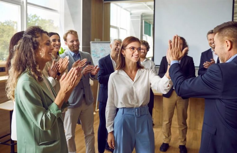 A group of people in business attire are clapping in the workplace as two individuals in the foreground high-five each other. The bright office setting features a whiteboard in the background, adding to the professional atmosphere.