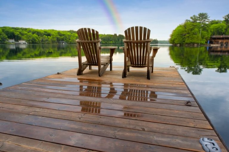 Two wooden chairs sit on a wet dock overlooking a calm lake, framed by lush green trees. The sky is clear with a faint rainbow reflected in the water, creating a serene, natural setting—a peaceful contrast to the fast-paced world of government just beyond this tranquil escape.