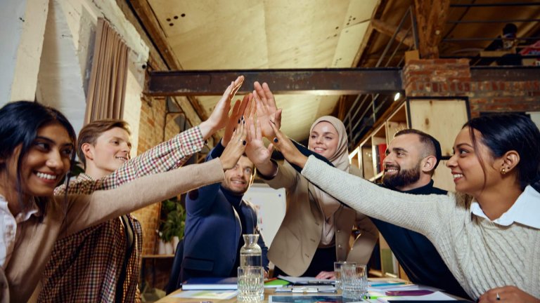 A diverse group of six colleagues sits around a table in an industrial-style office, smiling and giving a group high-five, celebrating teamwork, unity, and their commitment to DEI.