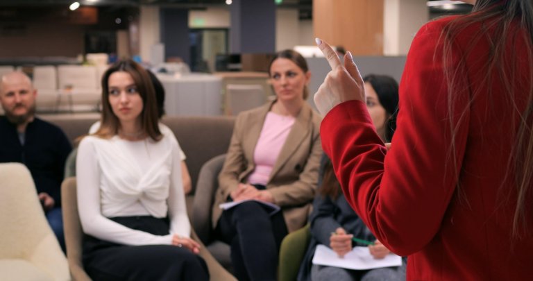 A person in a red blazer speaks to a seated group of four attentive adults—likely employers—in an office setting, with notebooks visible in the audience's laps.