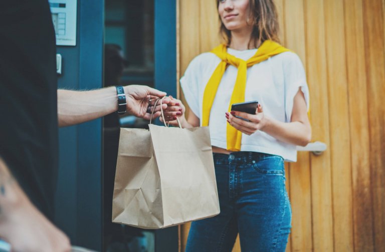 A gig worker hands a brown paper bag to a woman standing by a door. The woman, wearing a yellow sweater over her shoulders, holds a phone and is dressed in a white shirt and blue jeans.