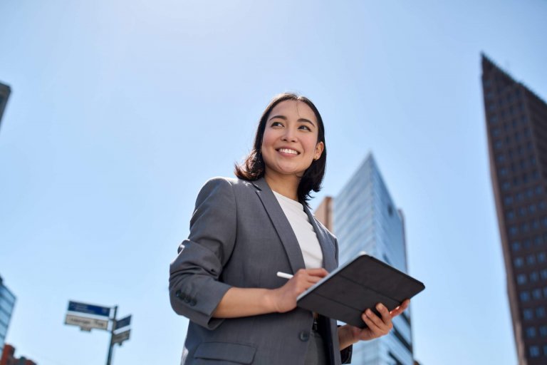 A woman in a gray blazer holds a tablet and smiles while standing outdoors in a city, ready for her job, with tall buildings and a clear blue sky in the background.