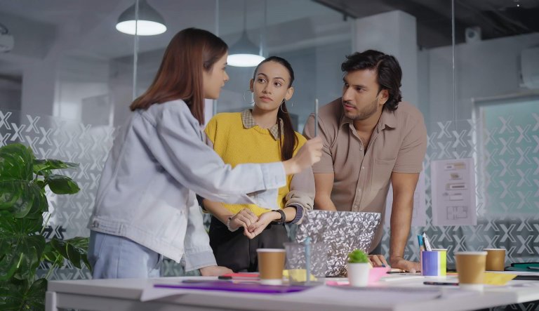 Three young adults stand around a desk in a modern office, engaged in a serious discussion about governments. Papers, coffee cups, and a laptop are on the table, with patterned glass walls in the background.