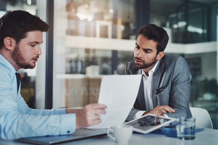 Two men in business attire sit at a table in an office, reviewing documents related to performance together. One holds a paper, while the other listens attentively. A tablet, notebook, and glass of water are on the table.