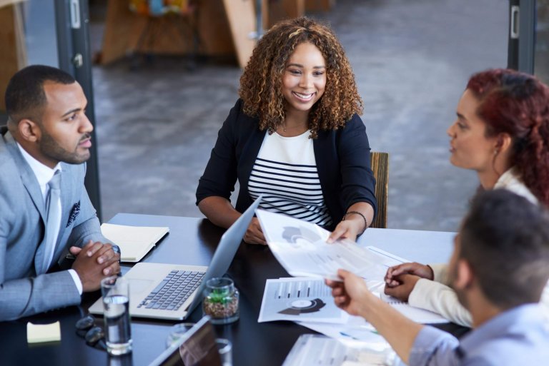 Four people sit around a table in a modern office, discussing documents and performance charts. One woman smiles while passing a paper to another, with laptops, papers, and glasses of water on the table.