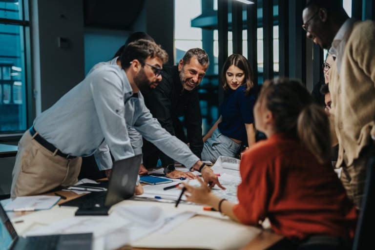 A group of colleagues gathers around a table covered with papers and laptops, engaged in a workplace discussion. One person points at a document while others listen attentively, suggesting teamwork and collaboration in an office setting.
