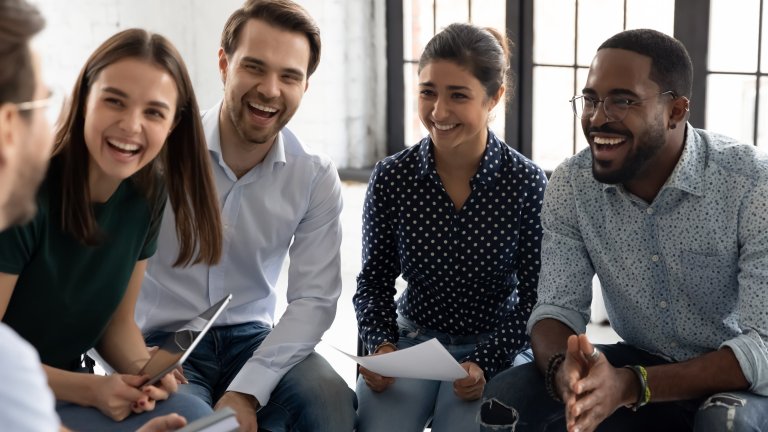 A diverse group of four talented young adults sit together indoors, smiling and laughing while holding papers, engaged in a lively conversation or discussion.