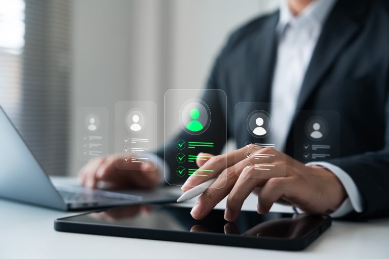 A person in a suit works on a laptop and tablet with a stylus. Floating digital icons show user profiles and checkmarks, suggesting public sector recruitment, selection, or digital HR management.