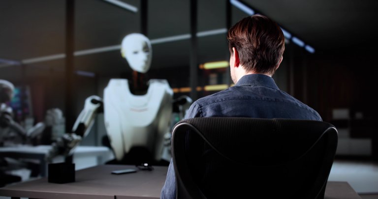 A person sits at a desk facing a humanoid robot across a table in a modern office setting, suggesting a conversation or interview between human and AI.