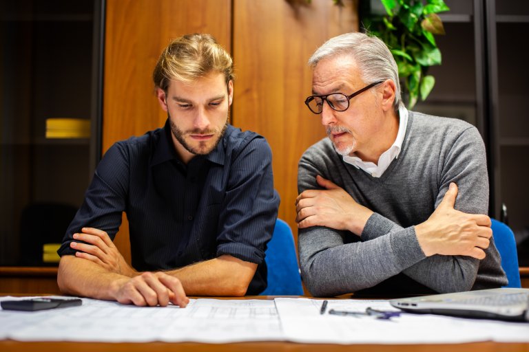 Two men sit at a workplace desk reviewing documents together. One man, with gray hair and glasses, looks at the papers while crossing his arms; the other points at the papers, both appearing focused.