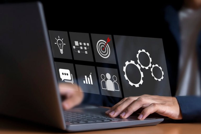 Hands typing on a laptop keyboard with digital icons floating above—including a light bulb, AI, flowchart, bullseye, chat bubble, bar chart, people, and gears—representing business innovation and technology concepts.
